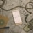 A flat lay composition featuring a smartphone with the word 'TRAVEL' displayed on its screen, positioned on top of several maps. A coiled piece of rope lies across the scene, accompanied by a wristwatch, a notebook with a pen, and some currency including paper bills and coins. The overall theme suggests preparation or planning for a journey.